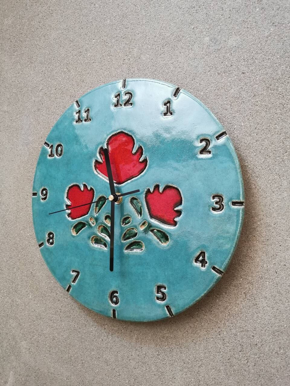 Zbliżenie na betonową ścianę, na której wisi zegar ceramiczny z ludowym motywem w kolorze niebieskim.