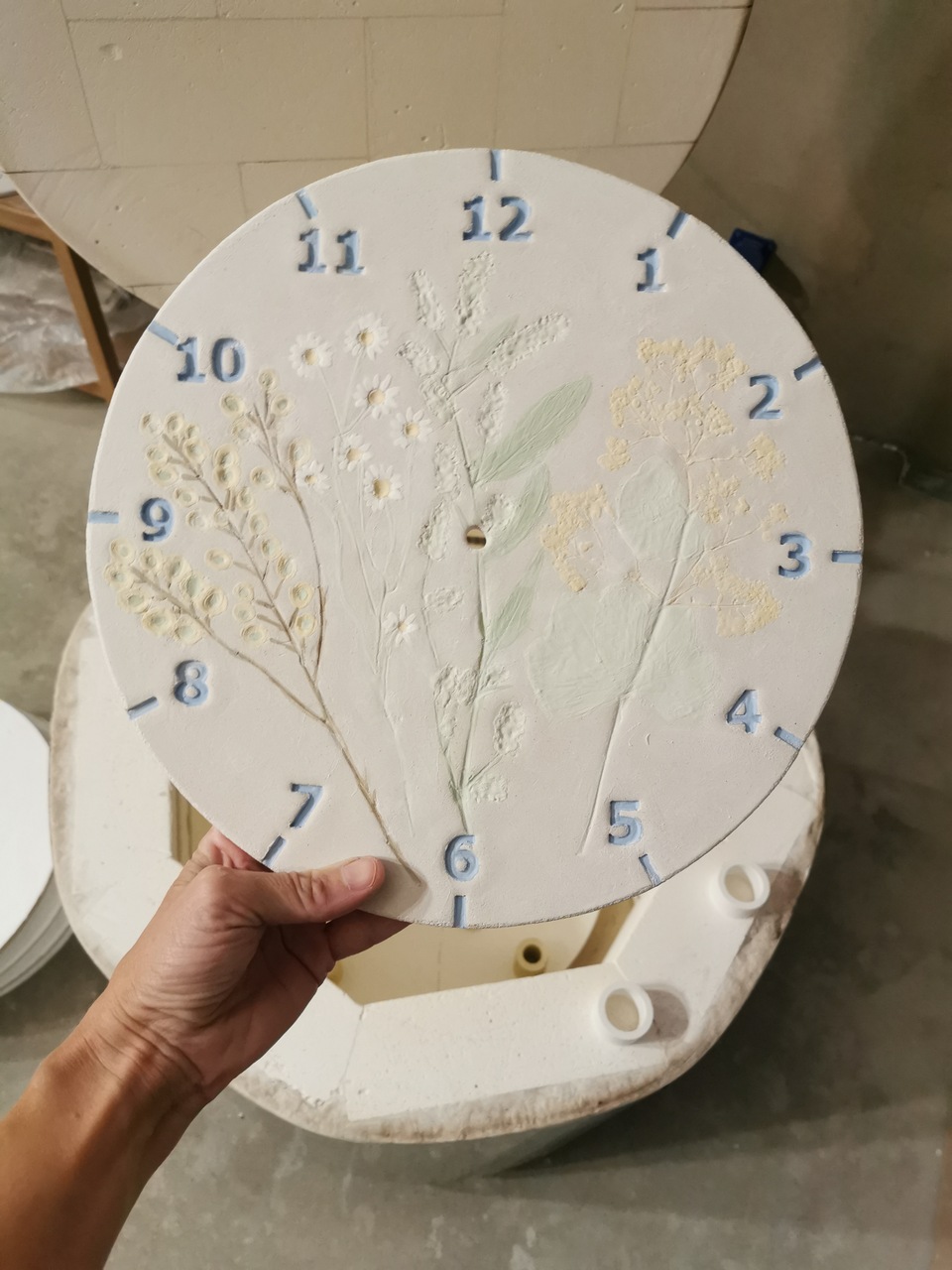 Ceramiczna tarcza zegara ręcznie malowana w trakcie powstawania w pracowni na tle ceramicznego pieca.